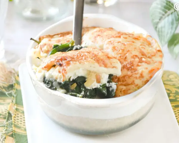 Deliciosa lasaña de espinacas gratinada, servida en un cuenco blanco. El queso dorado y burbujeante cubre las capas de espinacas y ricotta.