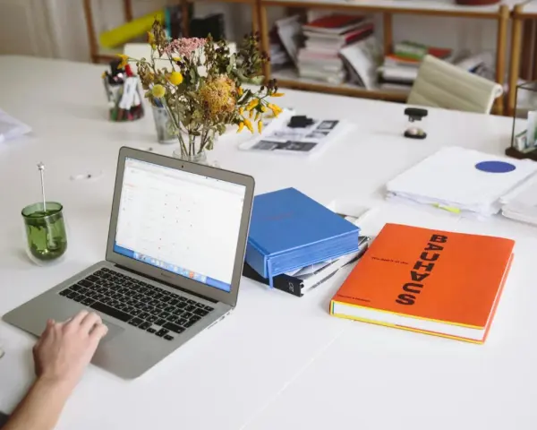 Escritorio con portátil, libros y flores. Un espacio de trabajo que parece una copistería por la cantidad de documentos y la organización.