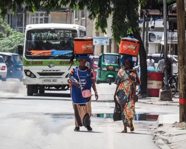 Dwie kobiety niosą na głowach pomarańczowe lodówki w upalny dzień w Dar es Salaam. W tle autobus i ruchliwa ulica.