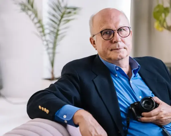 Tomaszewski fotograf w okularach, w granatowej marynarce i niebieskiej koszuli, trzyma aparat.