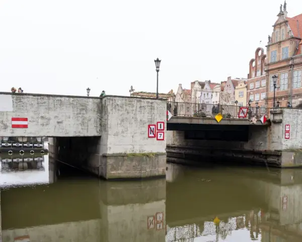 Nad kanałem w Gdańsku wznosi się betonowy zielony most, ozdobiony czerwono-białymi znakami. Ludzie spacerują po jego balustradzie.