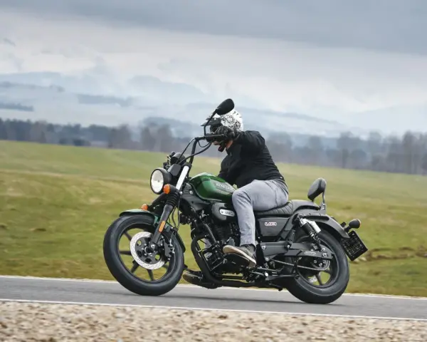 Motocyklista na zielonym chopperze UM Renegade 125 przemierza malowniczą drogę wśród zielonych pól i gór.