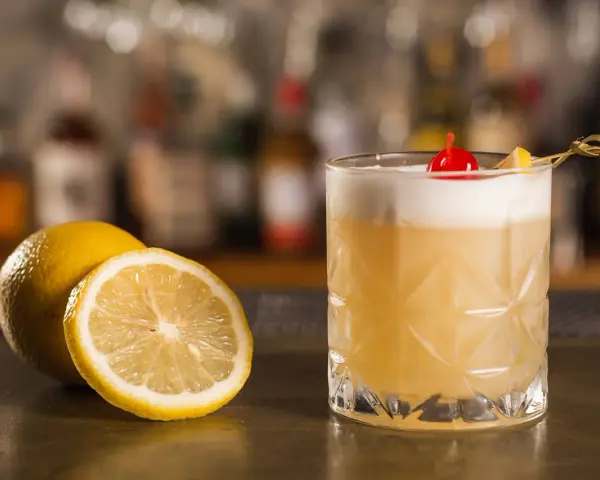 Orzeźwiający drink z amaretto, ozdobiony wiśnią i cytryną, stoi obok świeżych cytryn na barze.