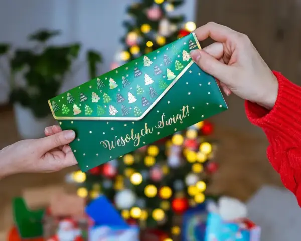 Jak zapakować voucher na prezent? Zielona koperta z choinkami i napisem "Wesołych Świąt!" jest przekazywana w świątecznej atmosferze.