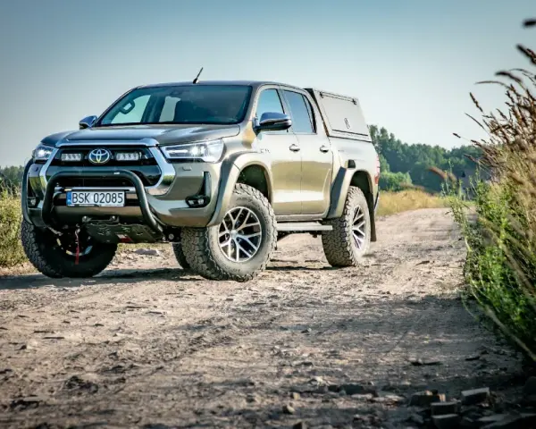 Złota toyota terenowa z wyciągarką i zabudową pędzi po polnej drodze, otoczona dziką roślinnością.