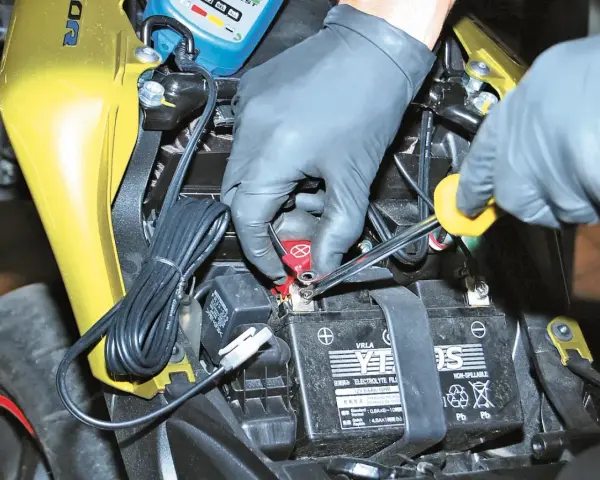 Mani guantate usano una chiave per staccare la batteria moto, scollegando il cavo rosso.