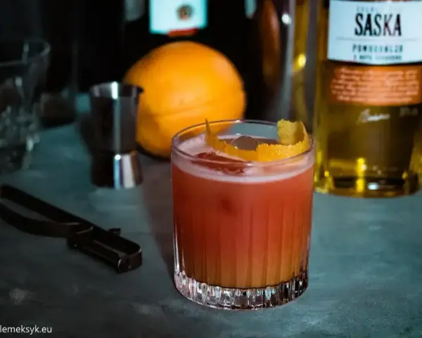 Orzeźwiający drink z likierem pomarańczowym, ozdobiony skórką cytrusową. Obok pomarańcza i butelka Saska.