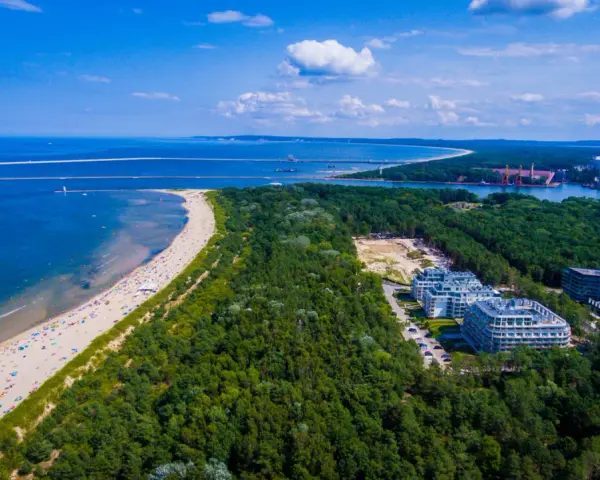 Widok z lotu ptaka na najszerszą plażę w Polsce, pełną wypoczywających ludzi, otoczoną lasem i nowoczesnymi budynkami.