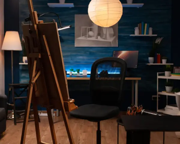 Un estudio de pintura en casa con caballete, silla de oficina, lámpara de pie y un escritorio con ordenador.