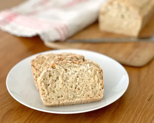 Skivor av hembakat bröd på en vit tallrik, perfekt för snabb bakning. En bit bröd ligger på en skärbräda i bakgrunden.
