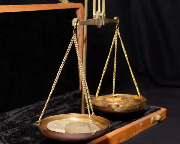 Złota waga szalkowa z monetami, gdzie jedna szalka zawiera kilka monet, a druga stosik krążków, symbolizując wagę uncja trojańska.