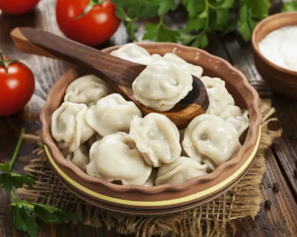 Pielmieni co to? Pyszne, gotowane pierożki w glinianej misce, z drewnianą łyżką, pomidorami i śmietaną w tle.