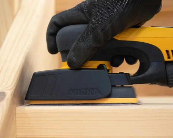 En hand med handske slipar en träyta med en Mirka DEOS-slipmaskin. Perfekt för att slipa borr och andra trädetaljer.