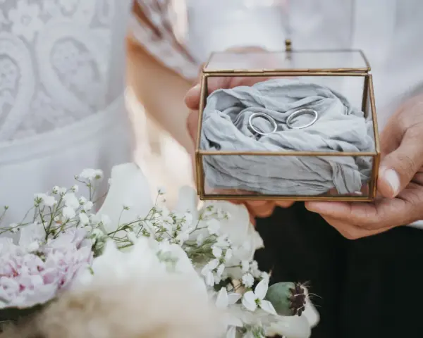 Dłonie trzymają szkatułkę z obrączkami, obok bukiet kwiatów. Niech te religijne życzenia na 50 rocznicę ślubu będą błogosławieństwem.
