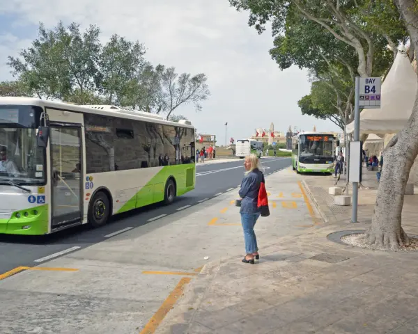 Kobieta czeka na autobus na Malcie. W tle widać autobusy, w tym jeden jadący w kierunku lotniska.