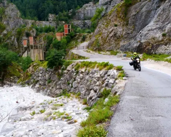 Un motociclista percorre una strada tortuosa tra le Alpi Apuane, con un fiume di sassi e ruderi industriali sullo sfondo.