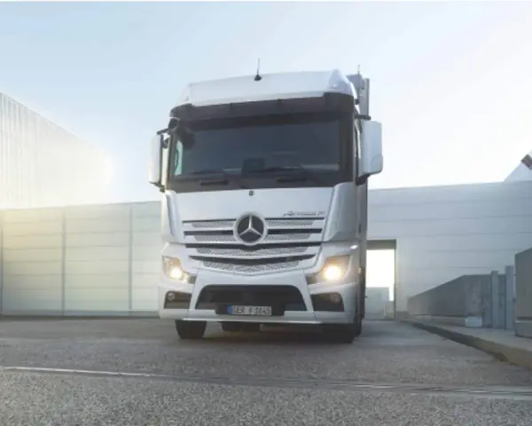 Biały Mercedes Actros MP4 z włączonymi światłami stoi przed nowoczesnym budynkiem.