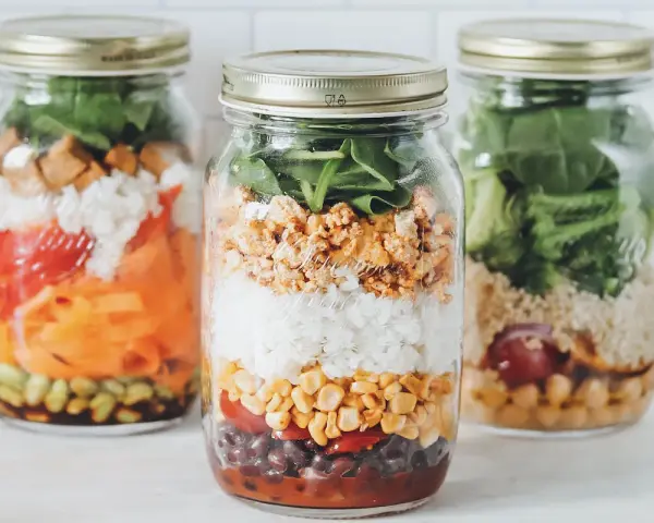 Drei bunte Schichtsalate im Glas, bereit für unterwegs. Jedes Glas ist ein kleines Kunstwerk mit Reis, Gemüse und Proteinen.