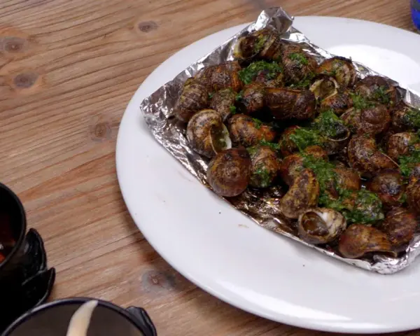 Caracoles con mantequilla y perejil, servidos en papel de aluminio sobre un plato blanco, junto a un cuenco con salsa.