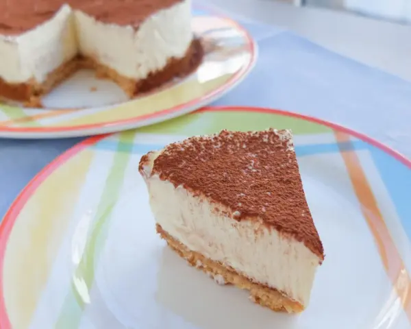 Una porción de tarta de queso fría, espolvoreada con cacao, reposa en un plato de colores. El resto de la tarta se ve al fondo.