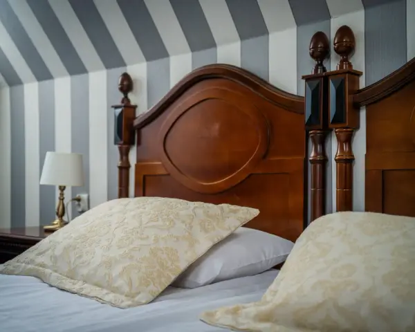 Eleganckie łóżko z drewnianym zagłówkiem i ozdobnymi poduszkami. Ciekawe, ile wynosi ryczałt za nocleg w takim miejscu?