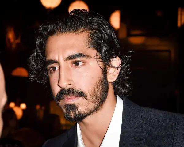 Primer plano de un hombre con cabello oscuro y rizado, y una barba poco poblada larga. Viste una camisa blanca y un traje oscuro.
