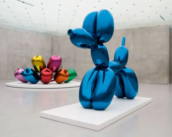 Un chien ballon bleu iconique de Jeff Koons, reflétant l'espace d'exposition. D'autres sculptures de ballons colorés sont visibles en arrière-plan.