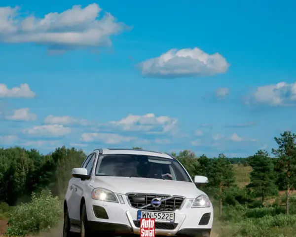 Białe Volvo XC60 T6 jedzie po piaszczystej drodze wśród zieleni, pod błękitnym niebem z chmurami.
