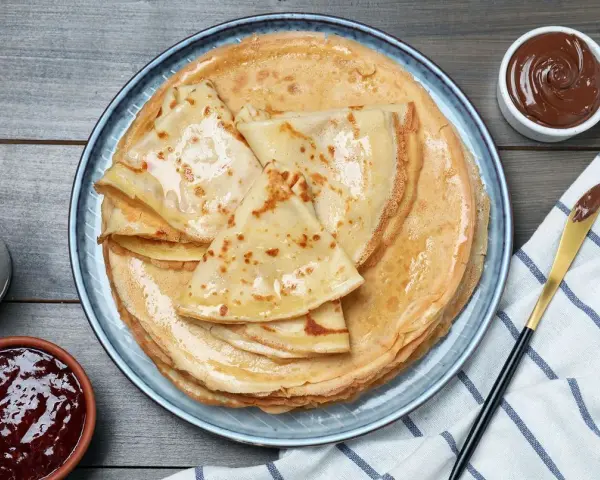 Pile de crêpes dorées, parfaites pour une recette pâte à crêpes. Accompagnées de confiture, pâte à tartiner et verre de lait.