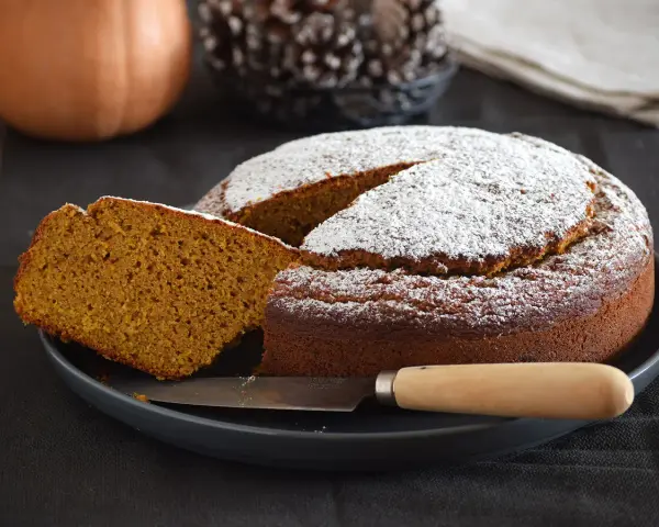 Bizcocho de calabaza esponjoso y dorado, espolvoreado con azúcar glas. Una rebanada muestra su interior húmedo y aromático.