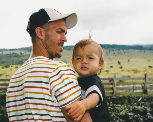 Ein Mann mit Bart und Kappe trägt ein Kind. Die Eigenschaften eines Papas zeigen sich in seiner liebevollen Umarmung.