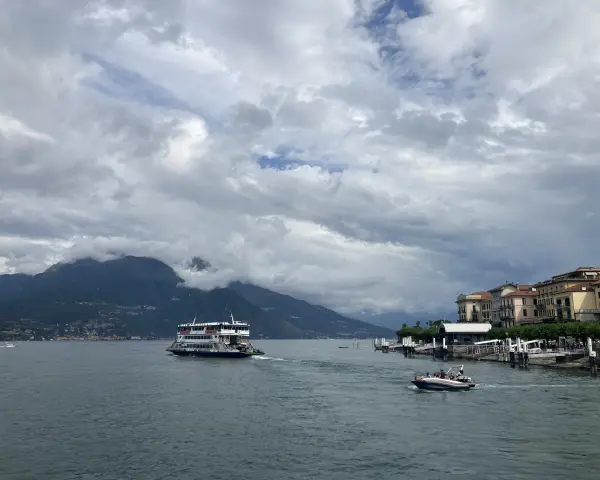 Promenada nad jeziorem Como, z górami w tle i statkiem wycieczkowym. W oddali widać urokliwe miasteczko.