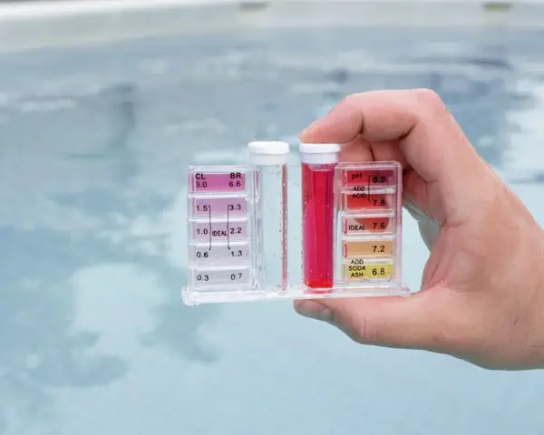 Hand holds pool water test kit. Red liquid in the pH tube indicates a high reading, suggesting acid is needed to lower pH.