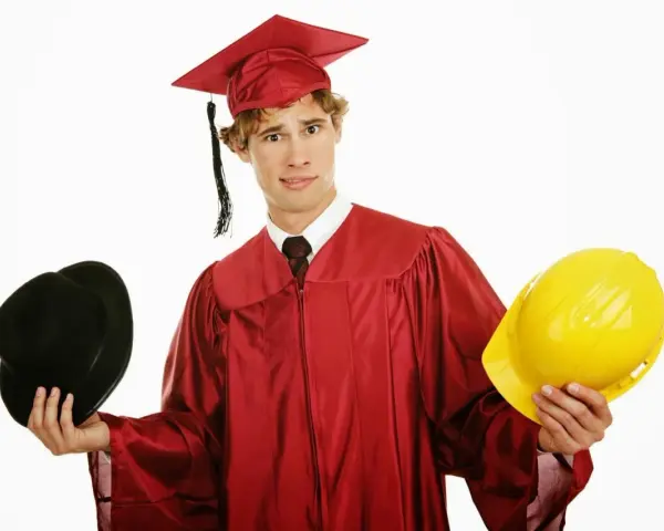 Studente universitario lavoratore con toga e cappello da laureato, tiene in mano un cappello nero e un casco giallo, indeciso sul futuro.