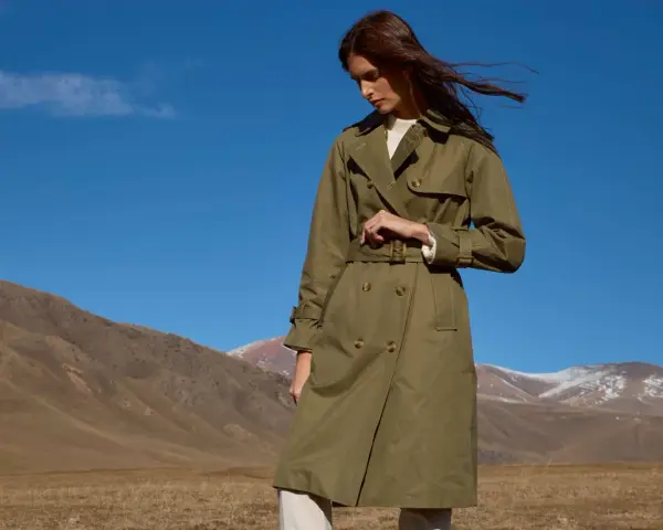 Mujer con un trench verde oliva, cinturón anudado, en un paisaje montañoso con cielo azul.