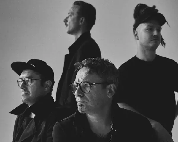 Czterech mężczyzn w czarnych ubraniach, spięty black metalowy zespół, z różnymi fryzurami i okularami, pozuje do zdjęcia.
