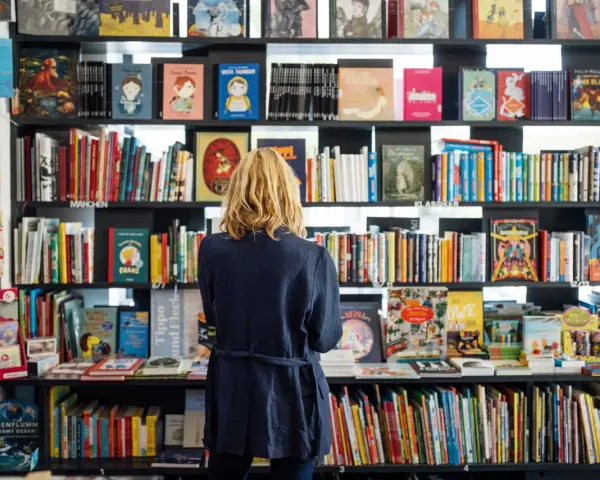 Donna bionda in libreria, circondata da scaffali pieni di libri colorati. Un'atmosfera di lavoro e scoperta tra le pagine.