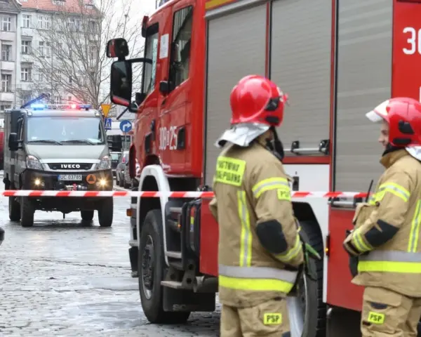 Dwóch strażaków w hełmach i mundurach stoi przy wozie strażackim. W tle wóz saperski. Starszy ogniomistrz zarobki to marzenie wielu, ale oni służą.