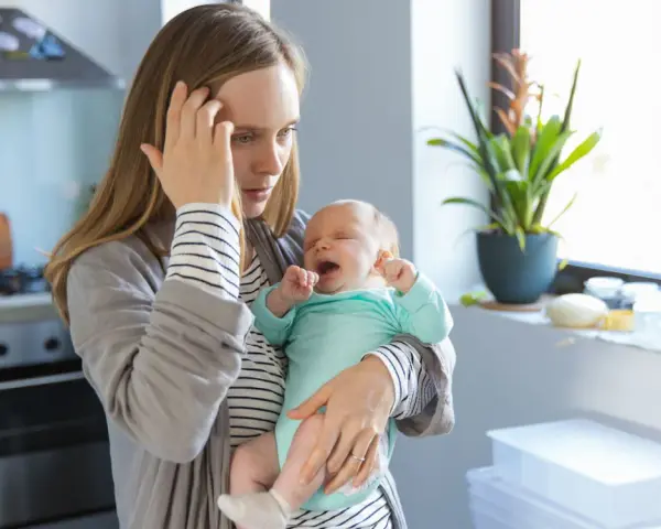 Müde Mutter hält weinendes Baby. Der **12 wochen schub** macht ihr zu schaffen. Sie hält sich die Stirn.