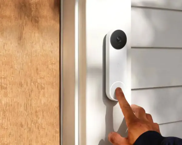 Un dedo presiona qué es el timbre inteligente, un dispositivo blanco con cámara negra, junto a una puerta de madera.