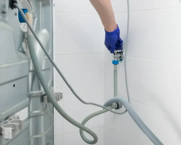 Blaue Handschuhe schließen den Wasserhahn für den Waschmaschinen Anschluss. Ein Schlauch führt zur Rückseite der Maschine.