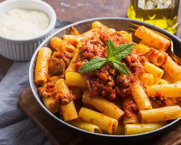 Un plato de macarrones con carne picada, cubiertos con salsa de tomate y decorados con menta. Al lado, un cuenco de queso rallado y una botella de aceite de oliva.