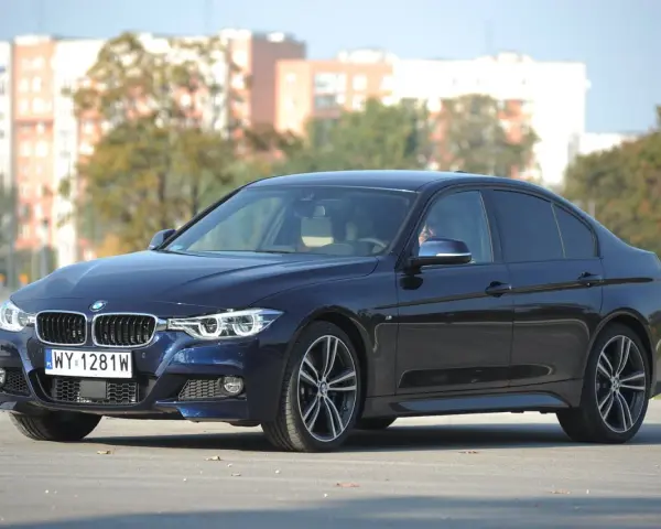 Ciemnoniebieskie BMW 318d z polskimi tablicami rejestracyjnymi stoi na tle budynków i drzew.
