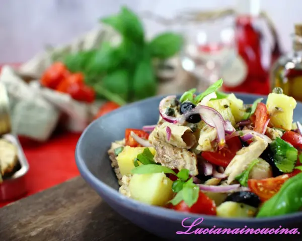 Un'insalata pantesca colorata con patate, pomodorini, olive, cipolla rossa e pesce, guarnita con basilico fresco.
