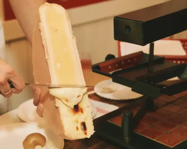 En person skrapar smält ost från en stor ostbit med en kniv. Perfekt för en mysig kväll med vin till raclette.