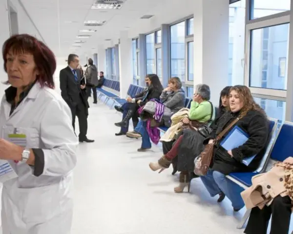 Doctora con bata blanca revisa papeles, mientras pacientes esperan en pasillo. Quizás pensando en cómo pedir excedencia estando de baja.