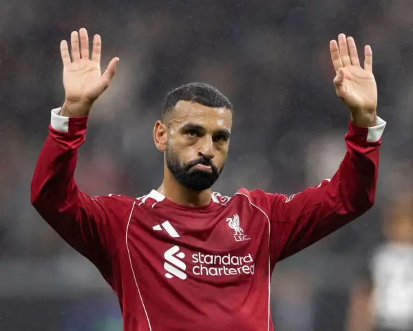 Mohamed Salah w czerwonej koszulce Liverpoolu, z uniesionymi dłońmi, pozdrawia kibiców. Widać logo klubu i sponsora.
