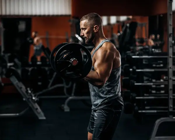 Mężczyzna ćwiczy biceps z ciężarem. W tle siłownia, sprzęt i inny ćwiczący.