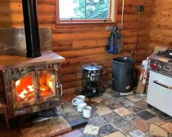 Poêle à bois allumé dans un chalet en bois rond, respectant la norme d'installation. Cuisine fonctionnelle avec cuisinière et ustensiles.