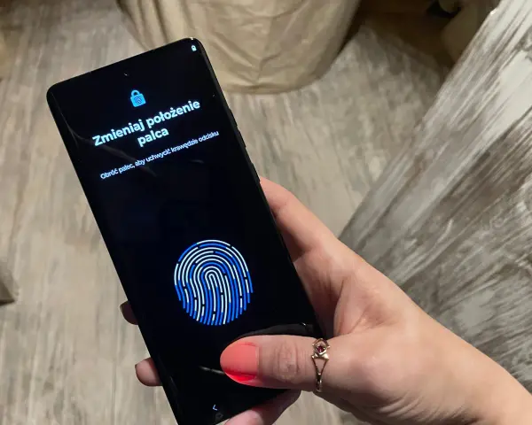 Dłoń z telefonem, na ekranie skaner linii papilarnych i komunikat "Zmieniaj położenie palca". To jak próba ucieczki z cyfrowego więzienia, niczym **jailbreak**.